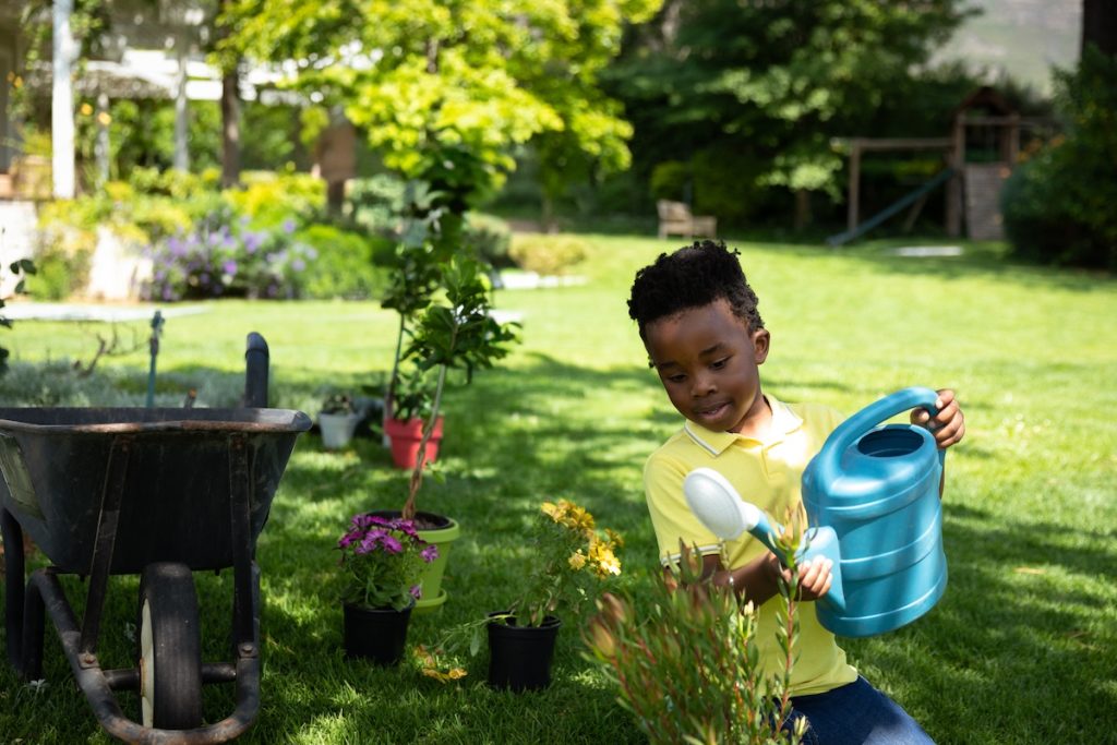 Un enfant en chemise jaune s'adonne joyeusement au jardinage enfant, arrosant les plantes avec un bidon bleu. À proximité, une brouette et des pots attendent, créant ainsi le décor idéal pour les activités de jardinage enfant.