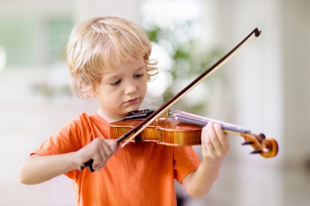 Un jeune enfant aux cheveux blonds vêtu d'une chemise orange joue du violon à l'intérieur, démontrant des compétences probablement perfectionnées en prenant des cours de musique.