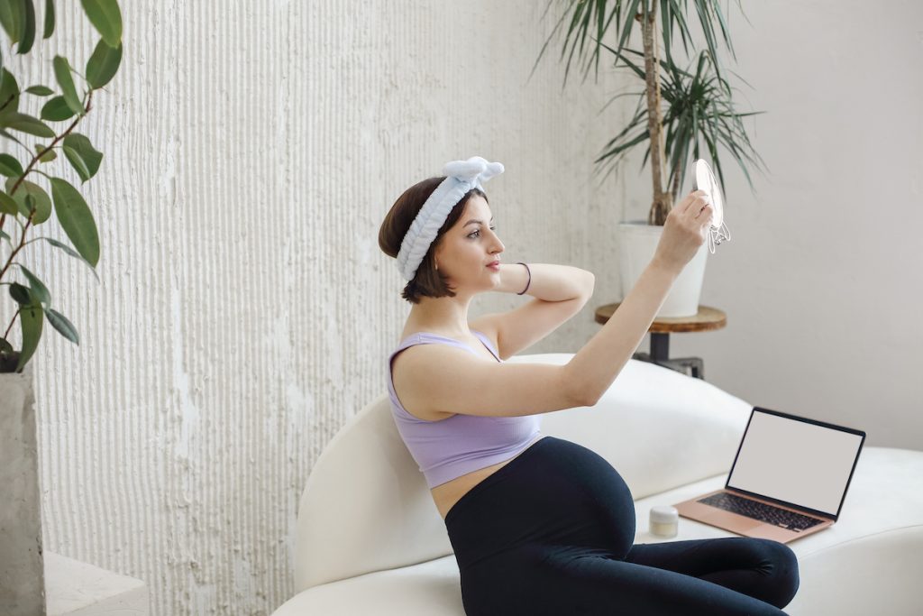 Une femme enceinte portant un bandeau et un pantalon de grossesse est assise sur un canapé dans ses vêtements de sport, prenant un selfie avec son smartphone. Un ordinateur portable et une plante sont en arrière-plan.
