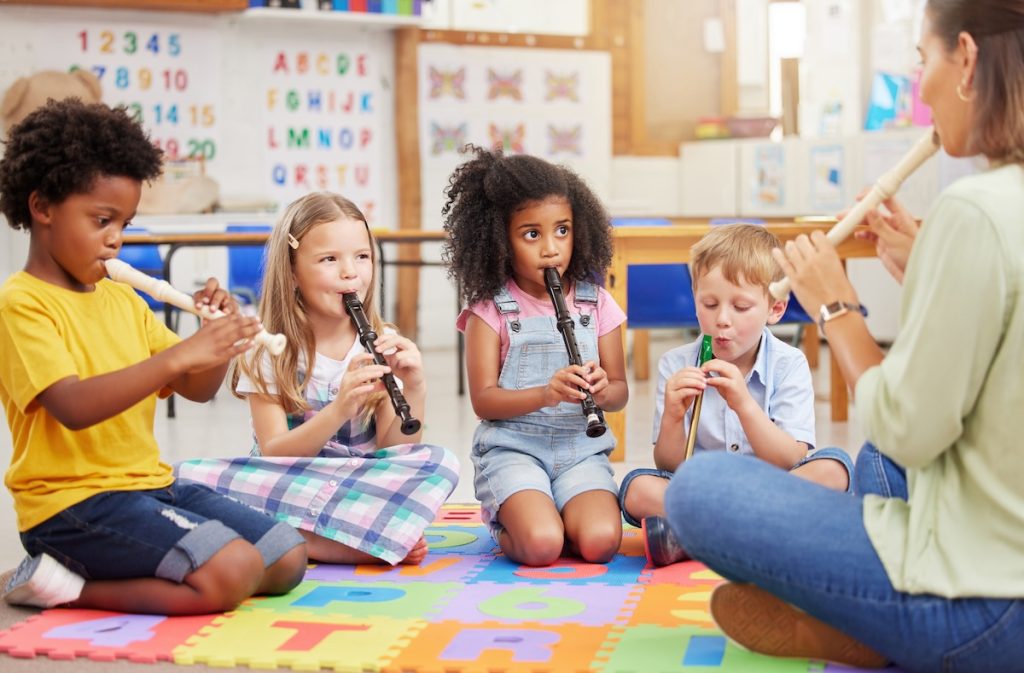 Un groupe d'enfants, assis sur un tapis coloré dans une salle de classe, participe à un cours de musique en jouant de la flûte à bec sous la direction d'un adulte.