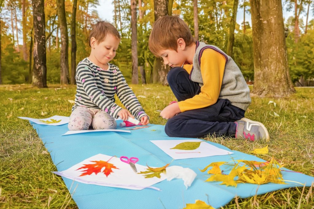 Deux enfants sont assis sur un tapis bleu à l’extérieur et participent à une activité de fabrication de feuilles avec du papier, des ciseaux et diverses feuilles colorées autour d’eux. Les arbres et la verdure sont en arrière-plan, créant un cadre idéal pour l'éveil des sens et l'apprentissage ludique.