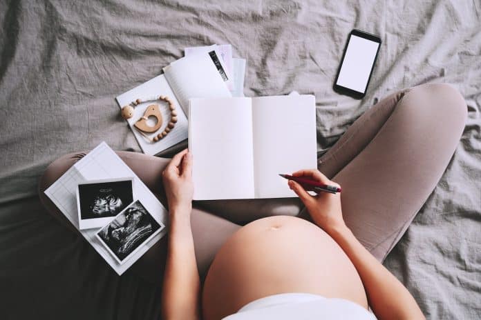 Une femme enceinte est assise sur un lit, mettant à jour sa liste de naissance sur son téléphone portable tout en prenant des notes dans un carnet pour l'arrivée de son deuxième bébé.
