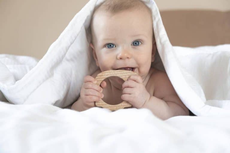Un bébé mâche un jouet en bois sous une couverture.