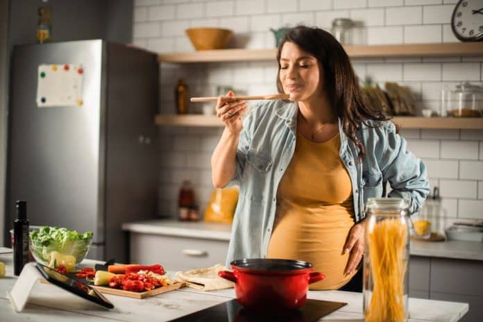 Une femme enceinte goûtant de la nourriture depuis une cuillère en bois dans une cuisine, avec des ingrédients et des épices pour la grossesse disposées sur