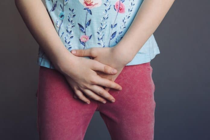 Un enfant vêtu d'une chemise à fleurs et d'un pantalon rose serrant les mains devant lui sur un fond gris, présentant éventuellement des symptômes nécessitant un diagnostic de pyélonéphrite.