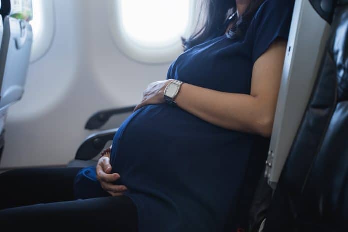Femme enceinte en chemise bleue prenant un vol près d'une fenêtre d'avion, tenant doucement son ventre.