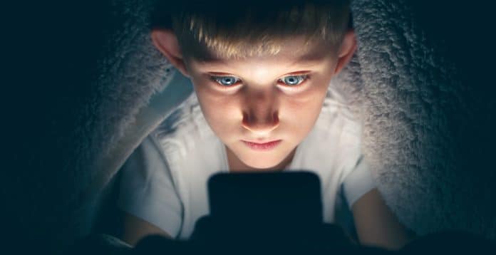 Un jeune garçon sous une couverture, éclairé par un écran lumineux, regardant intensément un smartphone dans une pièce sombre, inconscient de l'importance de la protection des données.