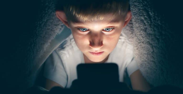 Un jeune garçon sous une couverture, éclairé par un écran lumineux, regardant intensément un smartphone dans une pièce sombre, inconscient de l'importance de la protection des données.