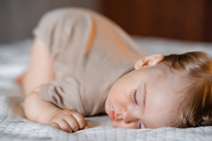 Un tout-petit aux cheveux châtain clair qui dort paisiblement sur un couvre-lit blanc texturé après un repas de rêve.