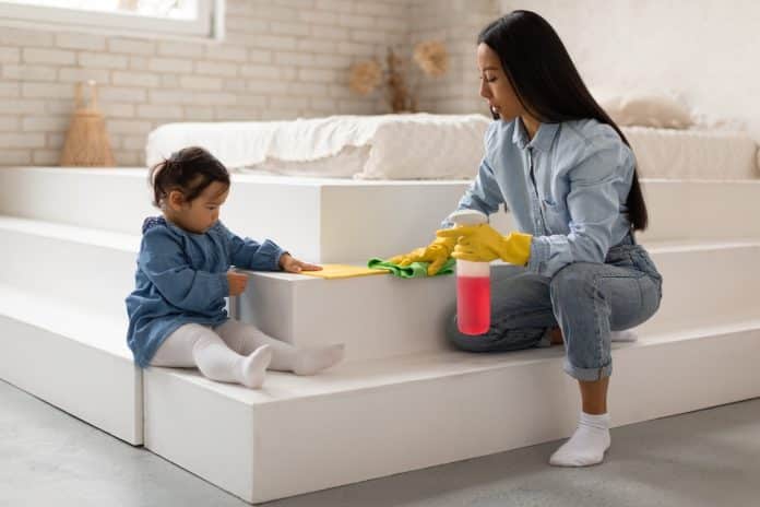 Une femme et un bébé en tenue de denim nettoyant un escalier blanc avec un chiffon jaune dans un salon lumineux.