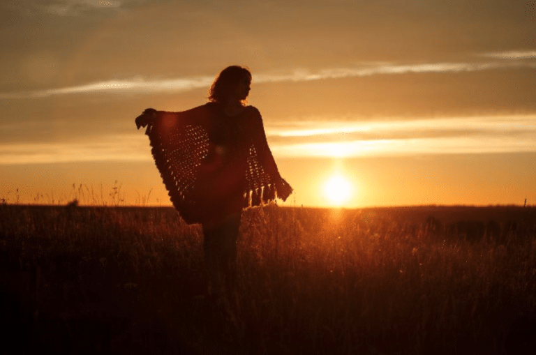 Silhouette d'une personne, peut-être un enfant, debout dans un champ herbeux au coucher du soleil, portant un poncho à pompons, avec le soleil bas à l'horizon.