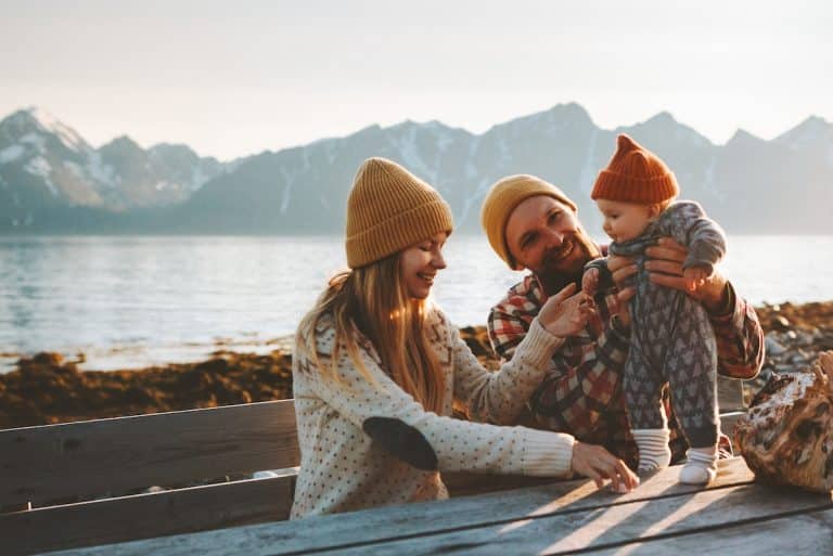 Un couple portant des chapeaux tricotés est assis sur un banc en bois au bord d'un lac avec des montagnes enneigées en arrière-plan, souriant et tenant leur bébé vêtu d'une tenue similaire, profitant au maximum de leur voyage avec bébé.