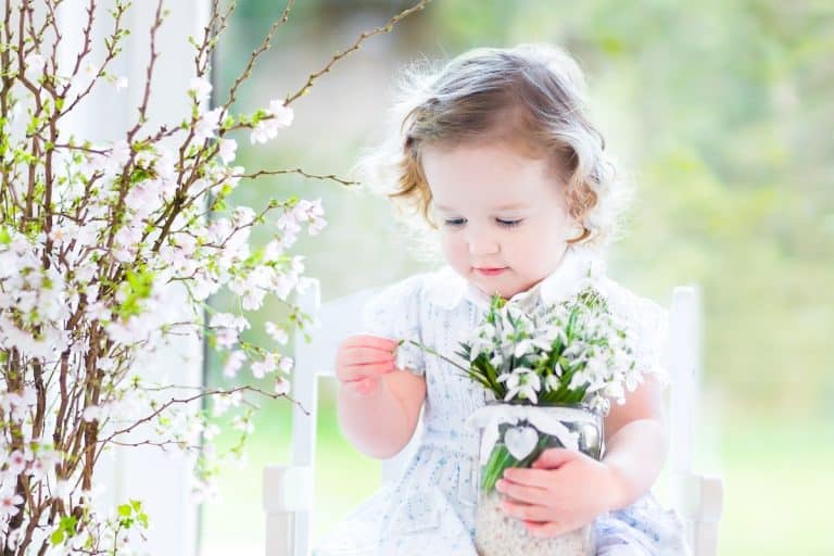 Un jeune enfant aux cheveux blonds bouclés tient un pot de fleurs blanches alors qu'il est assis sur une chaise blanche à côté d'une branche avec des fleurs blanches épanouies, une scène innocente qui semble être le parfait cadeau de baptême.