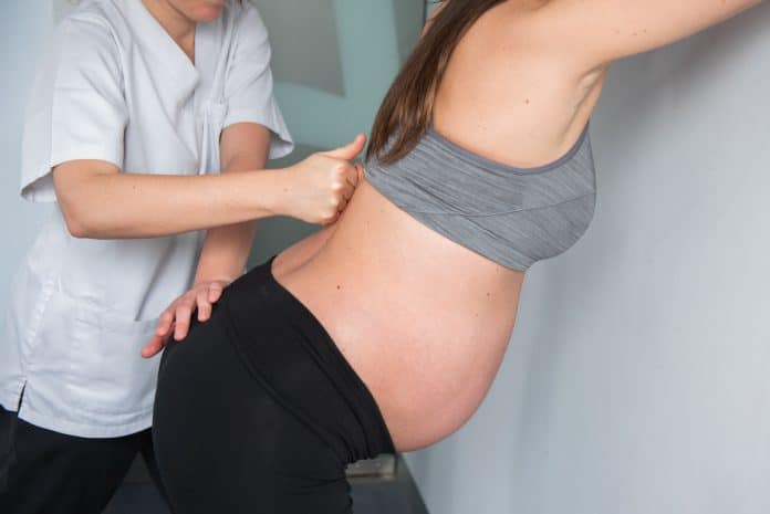 Une femme enceinte, se préparant à accoucher, reçoit un massage du dos de la part d'une personne en uniforme blanc. Elle est adossée à un mur, vêtue d'un soutien-gorge de sport gris et d'un pantalon noir.