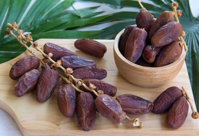 Un tas de dattes séchées sur une planche à découper en bois avec des dattes supplémentaires dans un petit bol en bois, placé sur une surface avec des feuilles vertes en arrière-plan : parfait pour celles qui se demandent si vous pouvez manger des dattes pendant la grossesse.