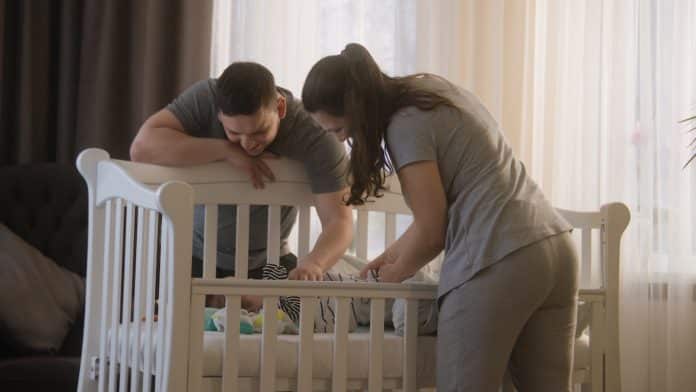 Deux adultes vêtus de vêtements gris assortis se penchent sur un berceau blanc et interagissent avec un bébé à l'intérieur, dans une pièce doucement éclairée et dotée de rideaux transparents. Leur attitude douce souligne leur engagement envers la sécurité sommeil de leur tout-petit.