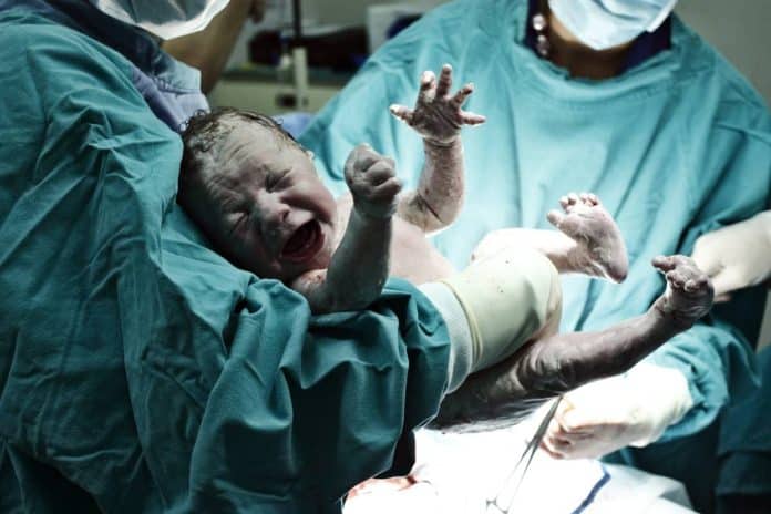 Un nouveau-né est tenu par un médecin dans une salle d'accouchement, couvert de vernix et pleurant, tandis qu'un autre membre de l'équipe médicale l'assiste. Les deux professionnels de la santé portent des masques chirurgicaux et des blouses.