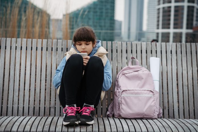 Une fille est assise sur un banc en bois et regarde un gobelet en papier qu'elle tient. Avec un sac à dos rose à côté d'elle et des bâtiments urbains flous en arrière-plan, elle semble inconsciente du monde trépidant des réseaux sociaux et de l'importance de protéger nos enfants de leur utilisation excessive.