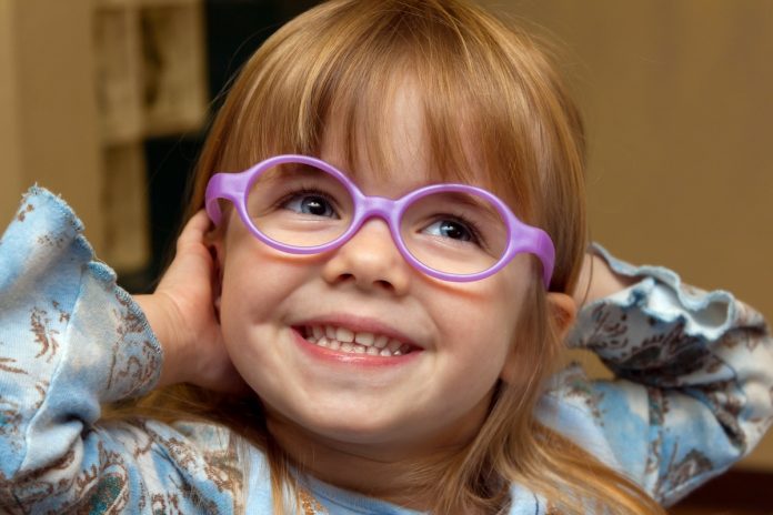 Un jeune enfant aux cheveux longs sourit largement en portant des lunettes violettes, une chemise bleu clair à motifs et en levant les mains près de la tête. Ce moment joyeux met en valeur l’innocence et l’énergie souvent observées chez les enfants, capturant un souvenir intemporel.
