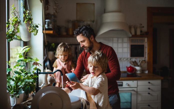 Un homme avec deux enfants faisant la vaisselle ensemble dans une cuisine présente des astuces pour impliquer les enfants dans les tâches ménagères. Des plantes sont suspendues près de la fenêtre et divers articles de cuisine sont visibles en arrière-plan.