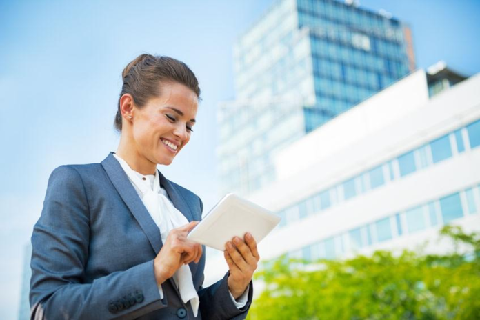 Une femme en tenue de travail se tient à l'extérieur devant un immeuble de bureaux moderne, souriant tout en utilisant des outils digitaux sur sa tablette.