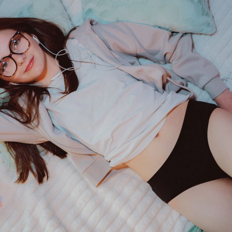 Une personne portant des lunettes, une chemise blanche et des sous-vêtements noirs est allongée sur un lit recouvert d'une couverture blanche et écoute des écouteurs.