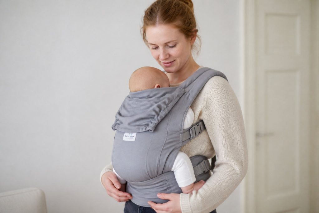 Une femme portant un pull beige porte un bébé dans un porte-bébé physiologique gris à l'intérieur, dans le cadre de notre sélection 2026 pour le meilleur porte-bébé.
