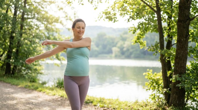 Une femme enceinte en tenue de sport, pratiquant un sport sans risque pendant son deuxième trimestre, étire ses bras en se tenant debout sur un sentier au bord d'un lac, entouré d'arbres et de verdure. Une femme enceinte en tenue de sport, pratiquant un sport sans risque pendant son deuxième trimestre, étire ses bras en se tenant debout sur un sentier au bord d'un lac, entouré d'arbres et de verdure.