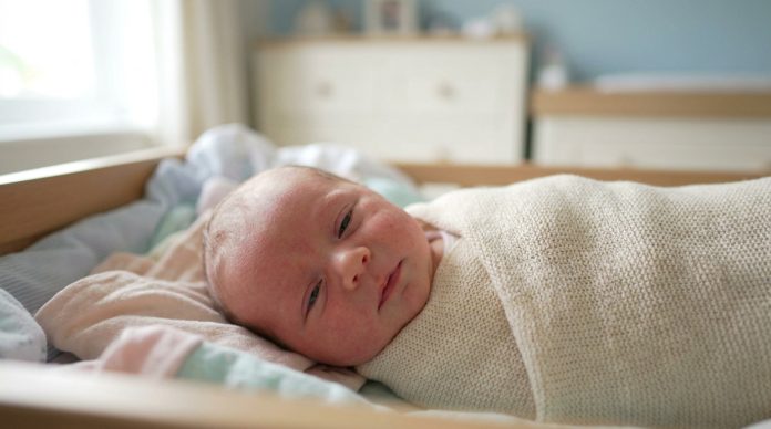 Un nouveau-né enveloppé dans une couverture beige est allongé sur une surface coussinée dans une pièce à l'éclairage tamisé et au mobilier blanc. Il présente des signes de fatigue bébé que les parents doivent reconnaître.