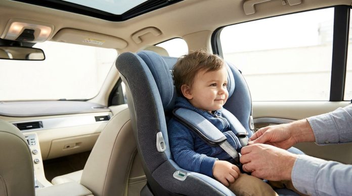 Un adulte attache un enfant souriant à l'aide d'un harnais dans un siège auto orienté vers l'arrière à l'intérieur du véhicule, garantissant ainsi la sécurité en fonction du poids de l'enfant.