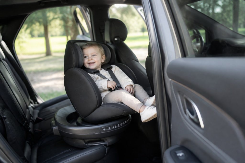 Un enfant souriant est assis dans un siège auto orienté vers l'arrière d'une voiture, vêtu d'un pull léger et d'un pantalon. La porte de la voiture est ouverte et des arbres sont visibles à l'extérieur - choisissez toujours un siège auto adapté au poids de l'enfant pour plus de sécurité.