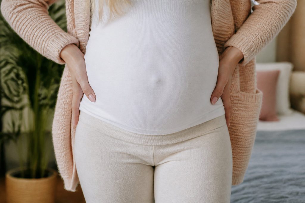 Une personne portant une chemise blanche et un pantalon beige se tient à l'intérieur, les mains sur les hanches, montrant un ventre de femme enceinte, essayant peut-être de soulager douleurs ligamentaires grossesse.